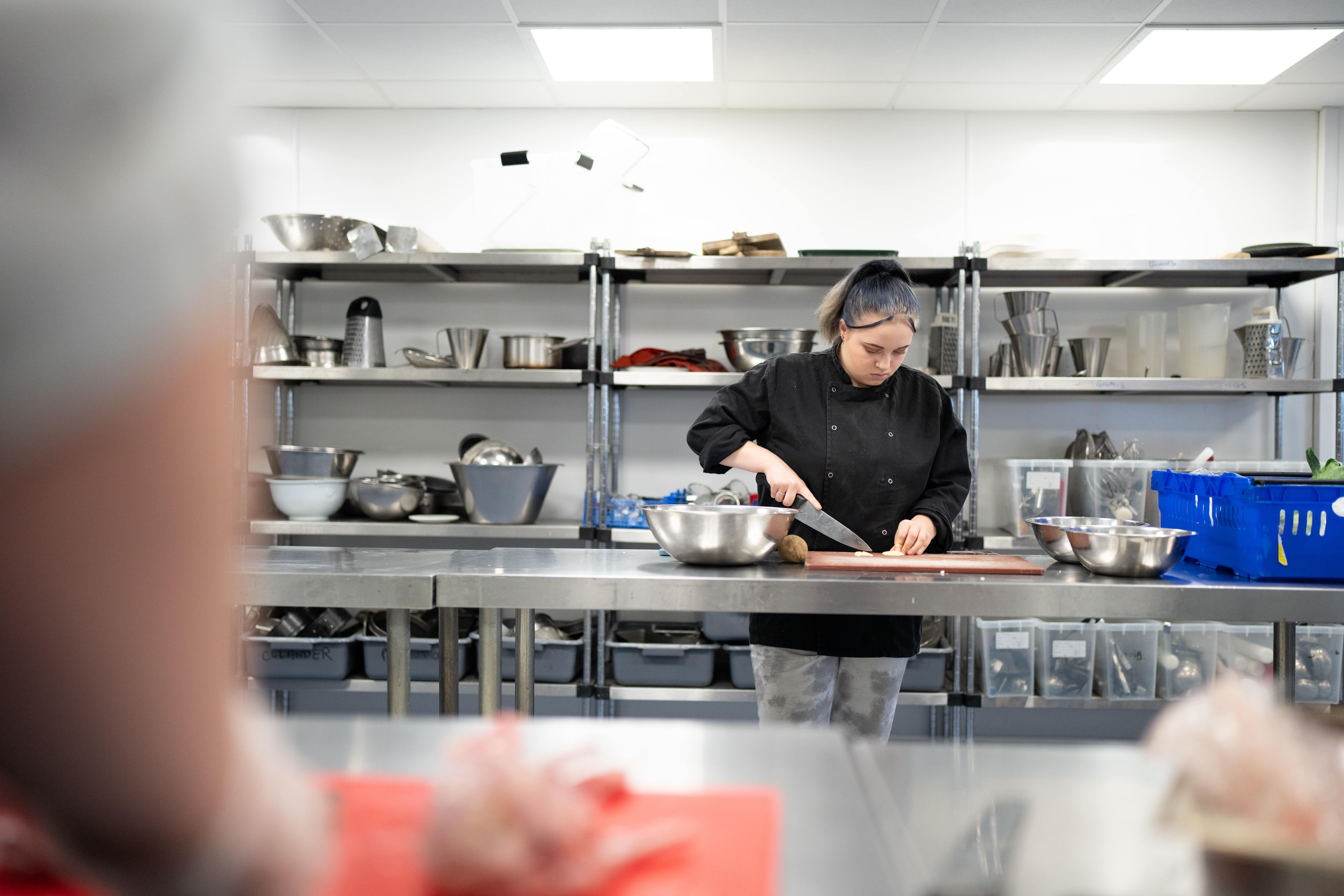 student chopping potatoes in training kitchen with shelves of cooking equipment in the background