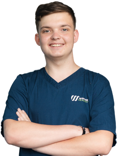 Young male healthcare student in blue medical scrubs with arms crossed, smiling confidently. The uniform features the 'Weston College Health' logo.