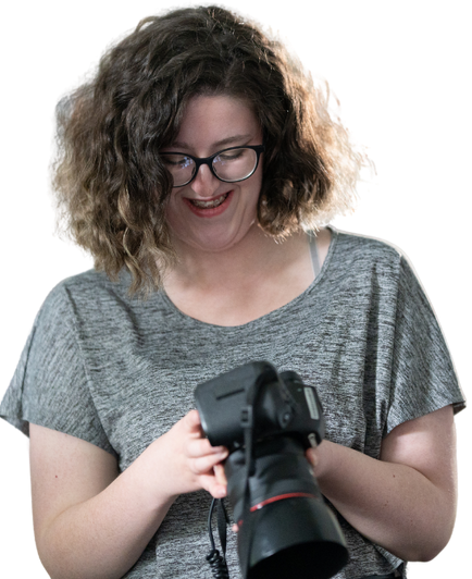 Smiling woman with curly hair and glasses looking down at a DSLR camera in her hands, wearing a gray shirt.
