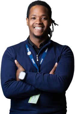 Smiling man with short dreadlocks, wearing a navy sweater, Airbus ID badge, and smartwatch, standing with arms crossed.