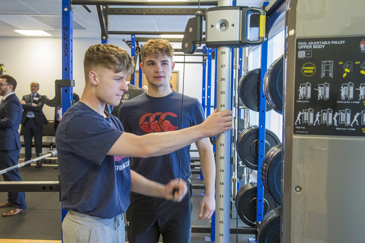 2 boys, one boy uses a gym cable machine while the other watches